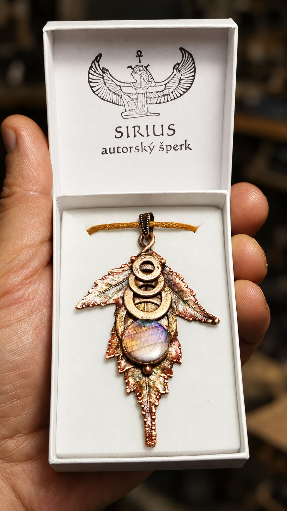 Měděný talisman Otisk podzimu s labradoritem v bílé krabičce s logem Sirius autorský šperk, držené v ruce.